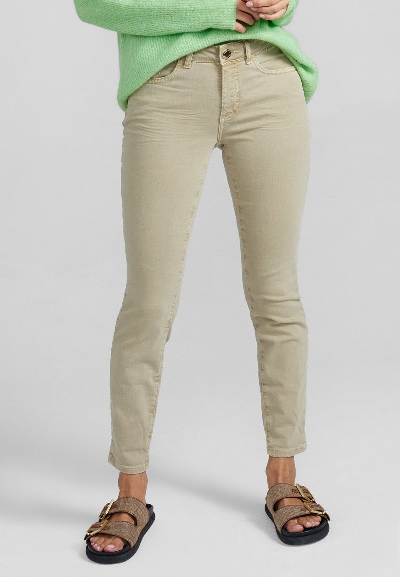 Personne porte un pantalon beige slim-fit, un pull vert et des sandales marron avec deux grandes boucles dorées, debout devant un fond gris uni.