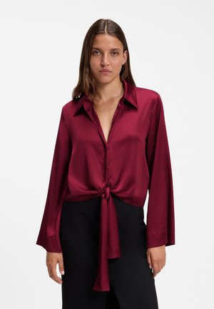 Eine Frau trägt eine langärmelige, tiefrote Satinbluse, die in der Taille gebunden ist, kombiniert mit schwarzen Hosen, vor einem schlichten weißen Hintergrund.