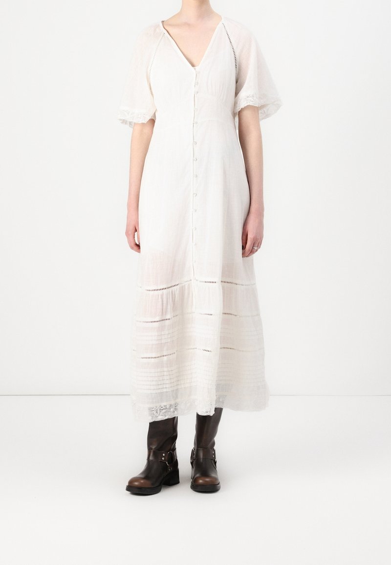 Robe midi blanche boutonnée avec manches bouffantes associée à des bottes en cuir noires mi-mollet sur un fond clair uni.