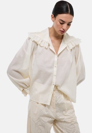 Blouse couleur crème avec un col à volants, des boutons sur le devant et des manches larges, associée à un pantalon léger à motifs avec des broderies complexes.