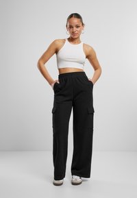 Schwarze Cargo-Hosen mit Seitentaschen, elastischem Bund und lockerer Passform, kombiniert mit einem weißen, verkürzten Tank-Top. Modell steht mit den Händen in den Taschen.