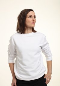 Frau mit schulterlangem braunem Haar, die ein weißes Langarmshirt und schwarze Hosen trägt, schaut zur Seite mit den Händen in die Hüften gestützt.