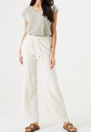 Kobieta ubrana w luźne, jasnobeżowe spodnie z troczkiem, jasnoszaro-brązowy krótki top z krótkim rękawem oraz brązowe sandały z odkrytymi palcami, stojąca na tle jednolitej białej ściany.