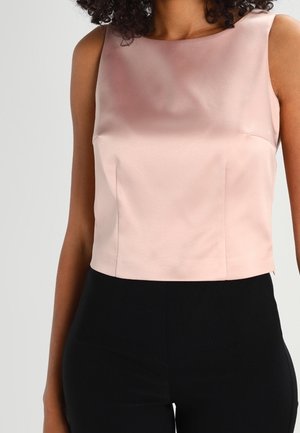 Femme portant un haut sans manches en satin rose clair, associé à un pantalon noir tailles hautes, sur un fond uni.