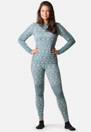 Donna in piedi con le mani sui fianchi, indossa un top a maniche lunghe e leggings abbinati con fantasia blu-grigio e calzini neri alla caviglia, su sfondo bianco.