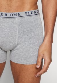 Boxers en coton gris avec une ceinture bleu marine, arborant le texte "PIER ONE." Vue rapprochée du vêtement avec une main sur la cuisse.