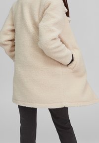 Abrigo de piel sintética color crema con un exterior texturizado, con bolsillos laterales y un dobladillo ligeramente acampanado, llevado sobre pantalones oscuros.
