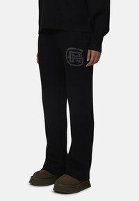 Czarne dzianinowe spodnie dresowe o luźnym kroju, z delikatnym szarym logo na lewej udzie. Tkanina ma gładką teksturę.