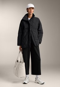 Zwarte gevoerde jas met een hoge kraag, gecombineerd met losse zwarte broek en sneakers. Houdt een lichtgrijze tote bag vast. Witte pet op het hoofd.