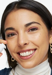 Person med medelhudton, axellångt mörkt hår, leende. Bär en vit polotröja, guldörhängen i-hoop, håller i en liten vit produkt.