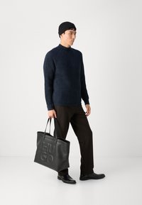 Maglione nero a coste, pantaloni marrone scuro, borsa a tracolla in pelle nera con logo "HUGO" impresso e scarpe nere, indossati con un berretto nero.
