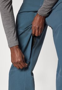 Ruke otvaraju bočnu zipersku džepu na plavim hlačama s vidljivom mrežastom postavom, koje su nosene preko sive majice s dugim rukavima, na jednobojnoj pozadini.