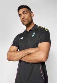 Black polo shirt with yellow stripes on the shoulders, turquoise logo accents, and pink side stitching, worn by a person with arms crossed.