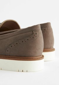 Scarpe in suede marrone con tacco basso, caratterizzate da un design perforato decorativo e una suola in gomma bianca, che mostra dettagli di texture e cuciture.