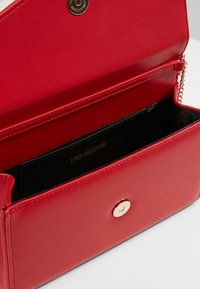 Červená peněženka z umělé kůže se zlatým uzávěrem, s černým interiérem a embosovaným logem. Kompaktní design s řetízkem.