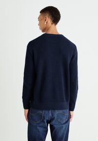 Tumši zili adīti džemperis ar teksturētu rakstu, apaļu kakla izgriezumu un ribinātu apakšu. Valkāts kopā ar tumši ziliem džinsiem, parādot ikdienišķu apģērbu no aizmugures.