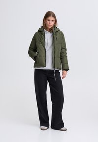 Olivgrüne Steppjacke mit Kapuze, Reißverschluss und Seitentaschen, getragen über einem grauen Hoodie, kombiniert mit schwarzen Jeans mit weitem Bein.