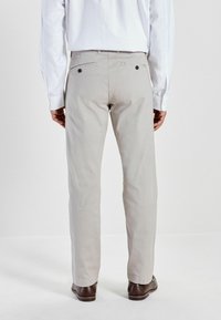 Pantalon ajusté gris clair avec une finition texturée, jambe droite, deux poches arrière avec des boutons noirs, porté avec une chemise blanche à manches longues.
