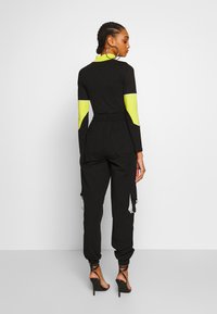 Svart jumpsuit med långa ärmar som har neongula detaljer på armarna, elastiskt midjeband och cargofickor. Högklackade skor fulländar looken.