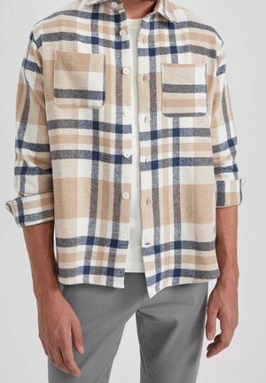 Mann trägt ein beige-weiß-navy kariertes Hemd mit Knöpfen, zwei Brusttaschen, hochgekrempelten Ärmeln, weißem Unterhemd und grauer Hose.