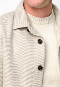 Giacca beige con motivo a spina di pesce e bottoni scuri, indossata sopra un maglione testurizzato color crema. Primo piano del colletto e dell'area del petto.