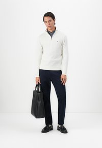 Mann i hvit genser med kvart glidelås, marineblå bukser, svarte loafers, som holder en svart skinn tote bag mot hvit bakgrunn.