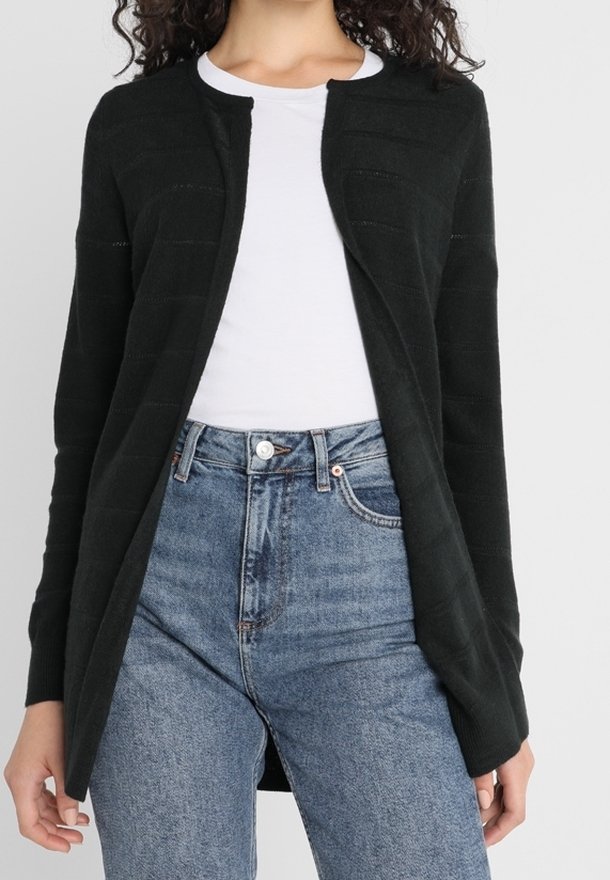 Kvinna klädd i en svart öppen cardigan över en vit t-shirt och högmidjade blå jeans, stående mot en enfärgad bakgrund.