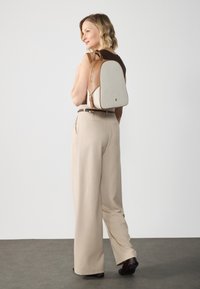 Mochila en tonos beige y marrón con una forma estructurada, cuenta con un cierre dorado y un acabado sutilmente texturizado. Llevada sobre los hombros con un atuendo casual.
