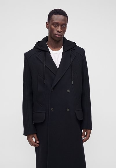Homme portant un manteau noir croisé avec un sweat à capuche en dessous, debout devant un fond gris clair uni.