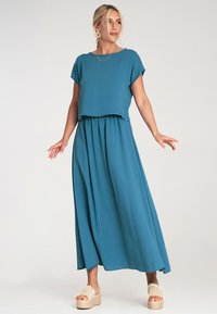 Turkusowy, krótki top z krótkimi rękawami, w połączeniu z luźną spódnicą maxi. Materiał ma miękką teksturę. Dobierany z jasnymi sandałami na platformie.