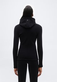 Άτομο που φοράει εφαρμοστή μαύρη φούτερ με κουκούλα και μαύρο παντελόνι, στέκεται με την πλάτη προς την κάμερα μπροστά σε απλό φωτεινό φόντο.