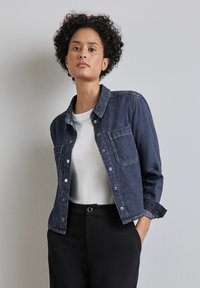 Kvinna med kort lockigt hår klädd i en mörkblå denimjacka över en vit skjorta och svarta byxor, stående mot en enfärgad ljus vägg.