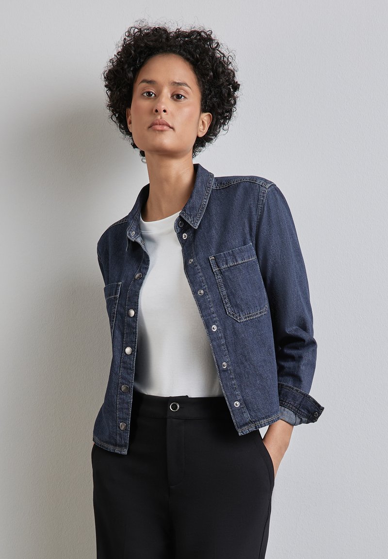 Kvinna med kort lockigt hår klädd i en mörkblå denimjacka över en vit skjorta och svarta byxor, stående mot en enfärgad ljus vägg.