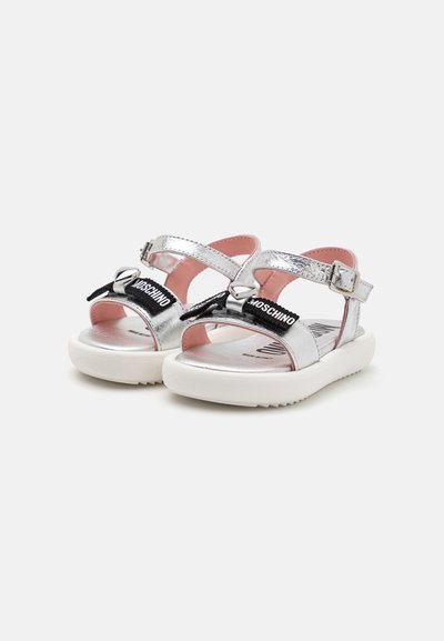 Sandales métalliques argentées avec une bride réglable, une marque noire et une semelle blanche rembourrée. Doublure intérieure rose pour le confort.