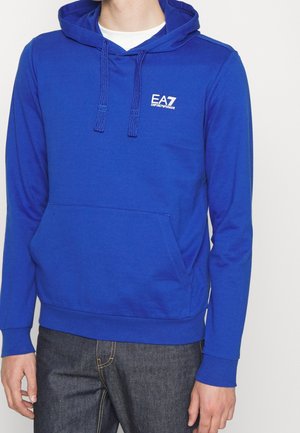 Mand iført en knaldblå EA7 Emporio Armani hættetrøje med forlommer og snøre, sammen med mørke denimjeans.