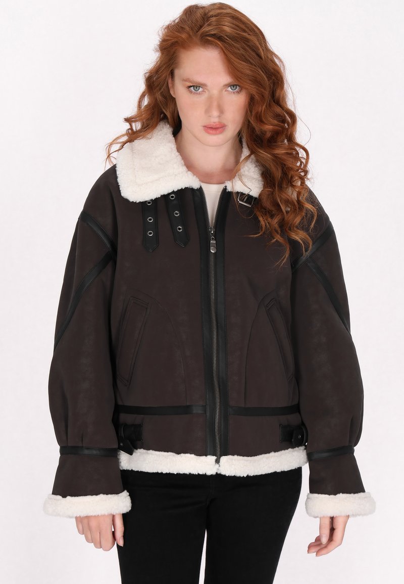 Ruskea keinonahkainen takki, jossa on valkoinen karvakaulus, vetoketjukiinnitys ja mustat yksityiskohdat. Takissa on ylipitkät hihat ja sivutaskut.