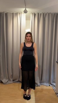 Žena stojící uvnitř na dřevěné podlaze, oblečená v černém tílku, průsvitné černé sukni a černých papučích, na pozadí šedé závěsy.