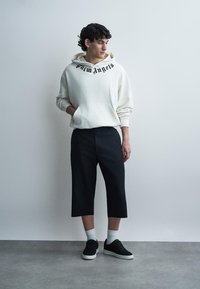 Mladi moški stoji z rokami v žepih, oblečen v belo kapucar majico "Palm Angels", črne široke kratke hlače, bele nogavice in črne superge.