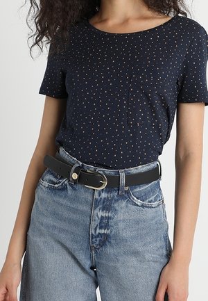 Mujer con una camisa de manga corta azul marino con lunares meterada dentro de unos vaqueros azul claro de talle alto con un cinturón negro.