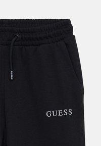 Čierne teplákové nohavice vyrobené z mäkkej látky, s elastickým pásom a bočnými vreckami. Názov značky "GUESS" je zobrazený v bielej výšivke.