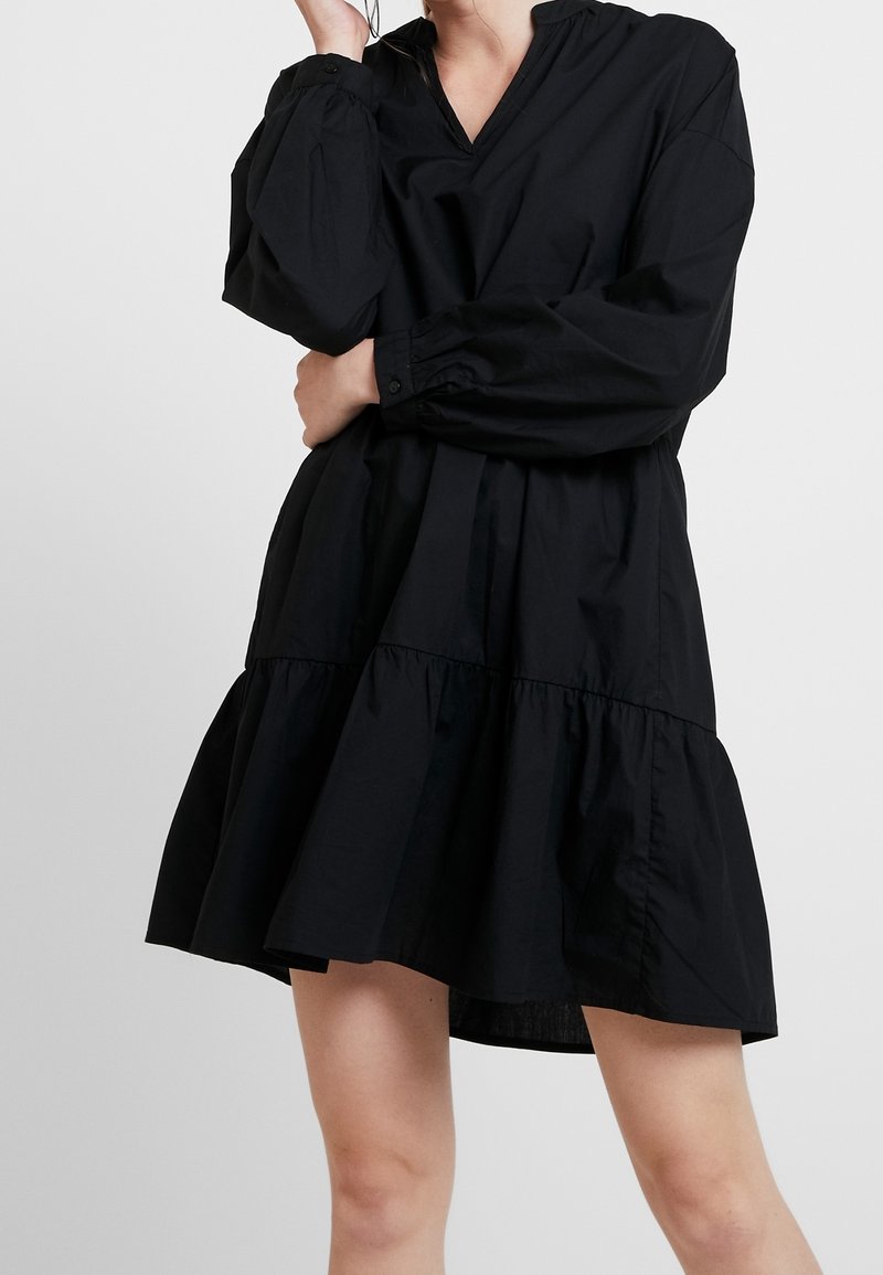 Femme portant une robe noire ample à manches longues avec une taille basse et un ourlet à volants, debout devant un fond uni.