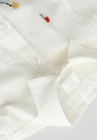 Chemise en coton blanc avec une finition texturée, dotée d'un col, d'une fermeture à boutons et d'accents brodés colorés près de l'épaule. Étiquette visible.