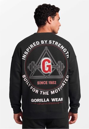 Man draagt een zwarte trui met een groot ontwerp op de rug waarop een halter in een driehoek en de tekst "Inspired by Strength, Built for the Motivated" te zien is.