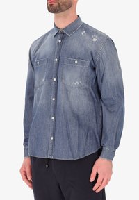 Camicia di denim di un blu chiaro, con due tasche sul petto, chiusura a bottoni e dettagli invecchiati sul tessuto. Maniche lunghe con polsini a bottoni.