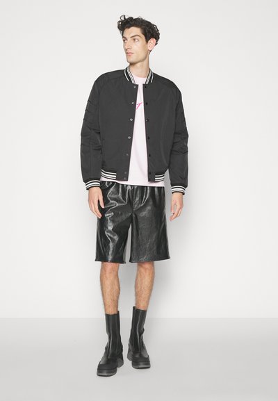 Svart bomberjakke med hvite striper på kragen og mansjettene, parret med blanke svarte skinnshorts og svarte ankelstøvler. Minimalistisk design.