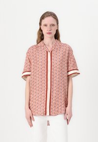 Kortearm, koraalkleurig shirt met een geometrisch patroon in donkerrood. Voorzien van een knoopsluiting aan de voorkant en een contrasterende witte streep op de mouwen.
