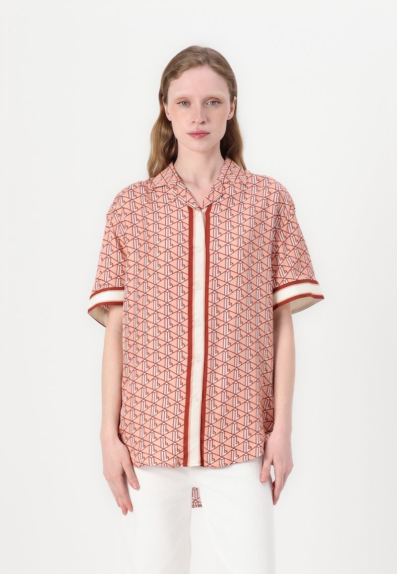 Kortearm, koraalkleurig shirt met een geometrisch patroon in donkerrood. Voorzien van een knoopsluiting aan de voorkant en een contrasterende witte streep op de mouwen.