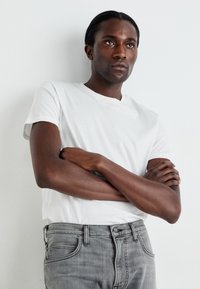 T-shirt blanc en coton avec un col en V, associé à un jean en denim gris à coupe classique et poches avant. Le modèle se tient devant un fond clair.