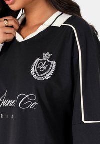 Vrouw in een zwart-wit geknoopte shirt met geborduurd embleem en de tekst "SA June, Co. Parijs" op de borst, hand bij de kraag.