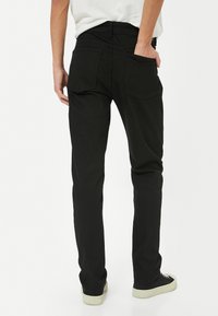 Vaqueros de mezclilla negros con corte ajustado, que cuentan con dos bolsillos traseros, pasadores para cinturón y una textura suave. Combinados con zapatos negros, vista lateral.
